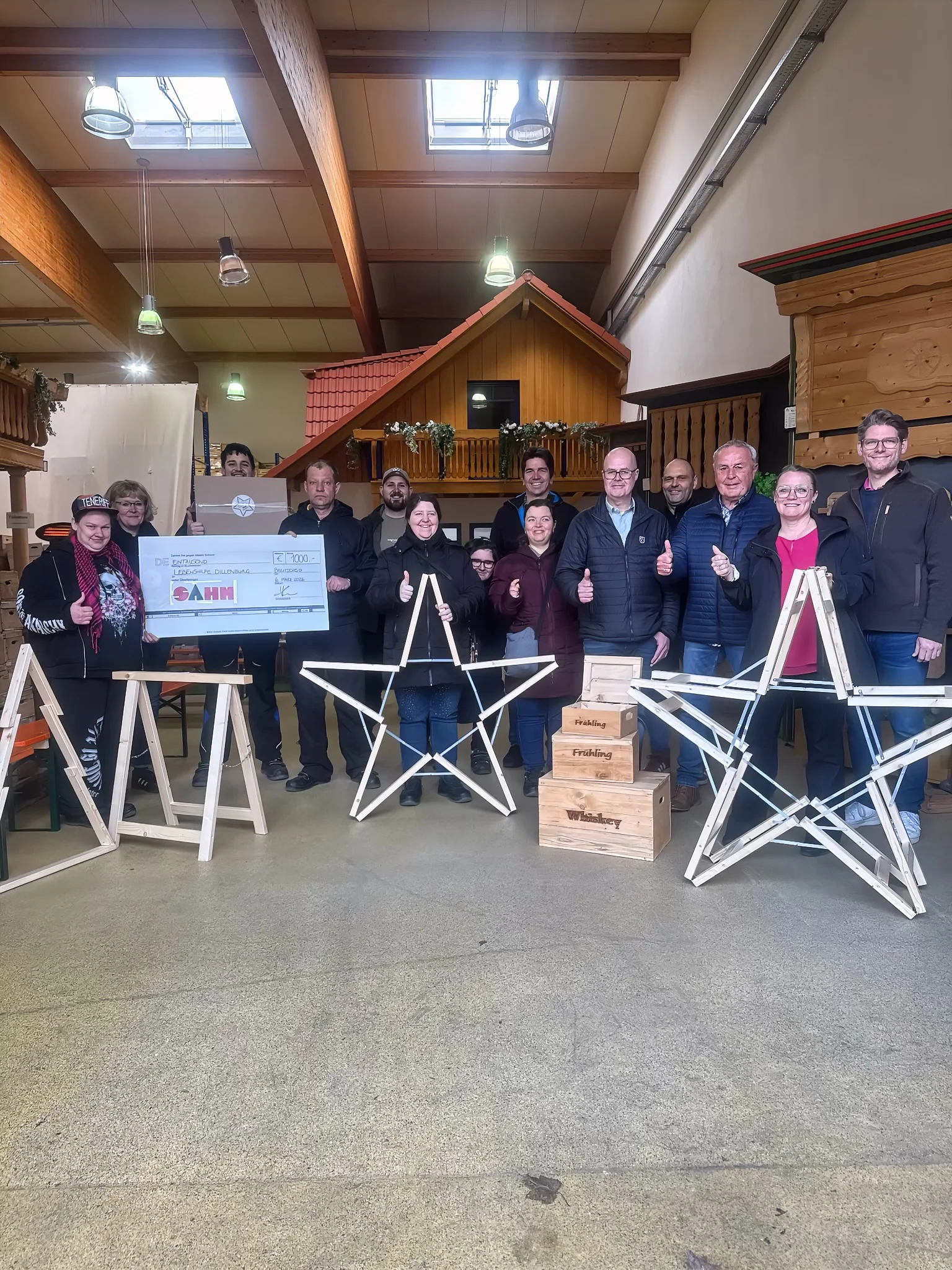 Eine Gruppe von Mitarbeitenden und Beschäftigten der Dillenburger Werkstätten sowie Vertreter der Firma Sahm stehen gemeinsam in einer Produktionshalle und halten symbolisch einen Spendenscheck über 1000 Euro hoch. Vor ihnen stehen mehrere große, aus Holz gefertigte Outdoor-Weihnachtssterne. Die Sterne sind das Ergebnis eines gemeinsamen Projekts zwischen der Sahm GmbH und den Dillenburger Werkstätten. Alle Beteiligten lächeln und zeigen Daumen nach oben.