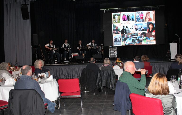Abendveranstaltung im Saal mit Liveband, gedeckten Tischen und Foto-Slideshow auf großer Leinwand.