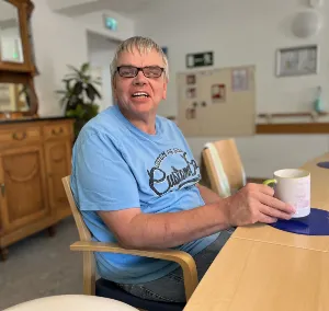 Älterer Mann lächelt mit Kaffeetasse am Holztisch im hellen Gemeinschaftsraum.