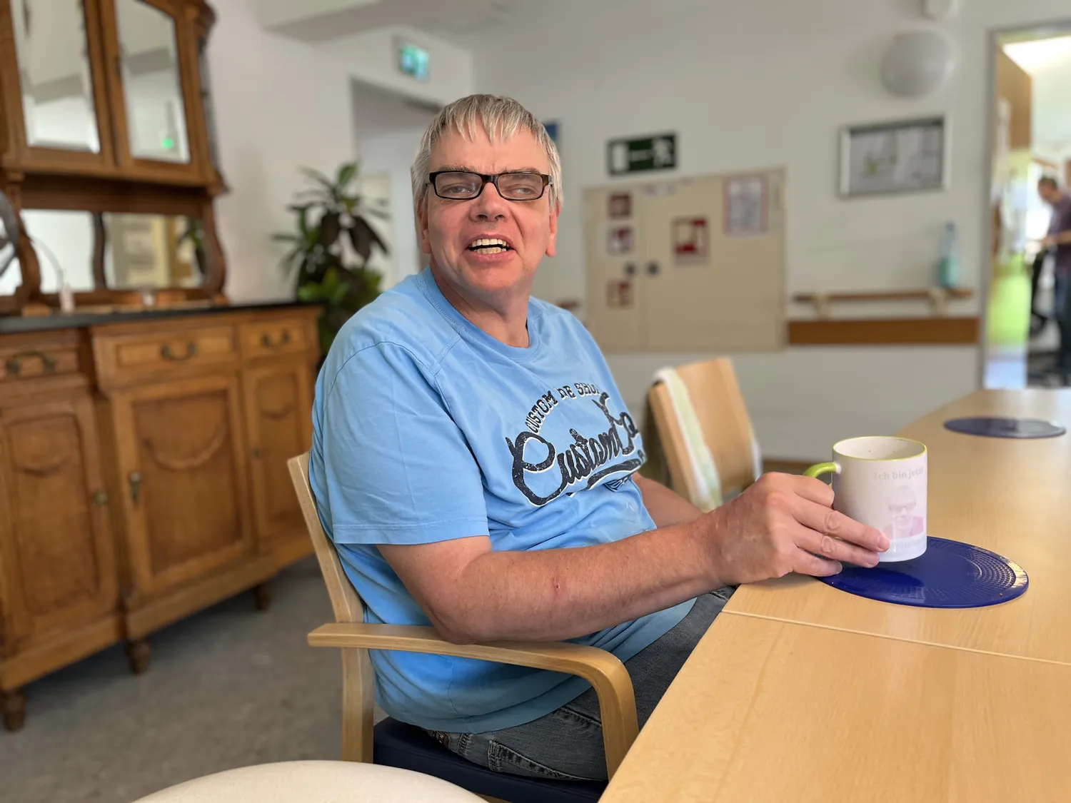 Fröhlicher Senior trinkt Kaffee im Gemeinschaftsraum, sitzt am Holztisch mit Tasse.