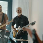 Fröhliche Bandprobe: Gitarrist und Bassist spielen, Sängerin mit Mikrofon im Proberaum.