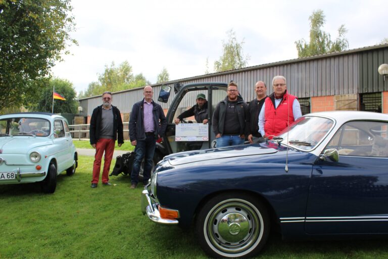 Sechs Männer bei Benefiz-Oldtimer-Treffen mit Scheck und klassischen Autos vor Werkstatt.