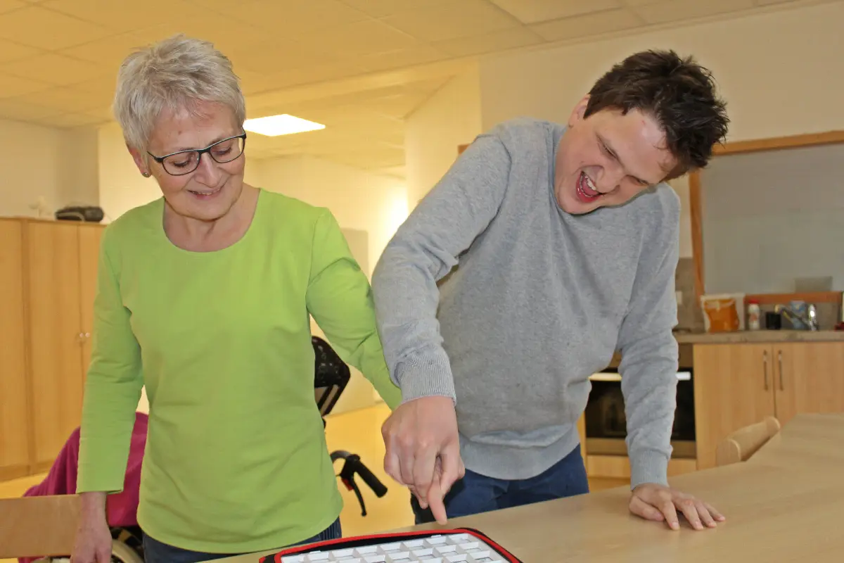 Betreuerin unterstützt Mann bei AAC-Kommunikation am Tisch, beide lachen gemeinsam.