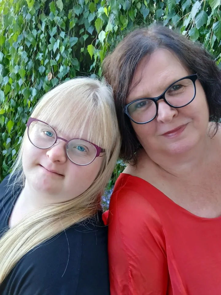 Zwei Frauen mit Brille lächeln vor grüner Pflanzenwand im Freien