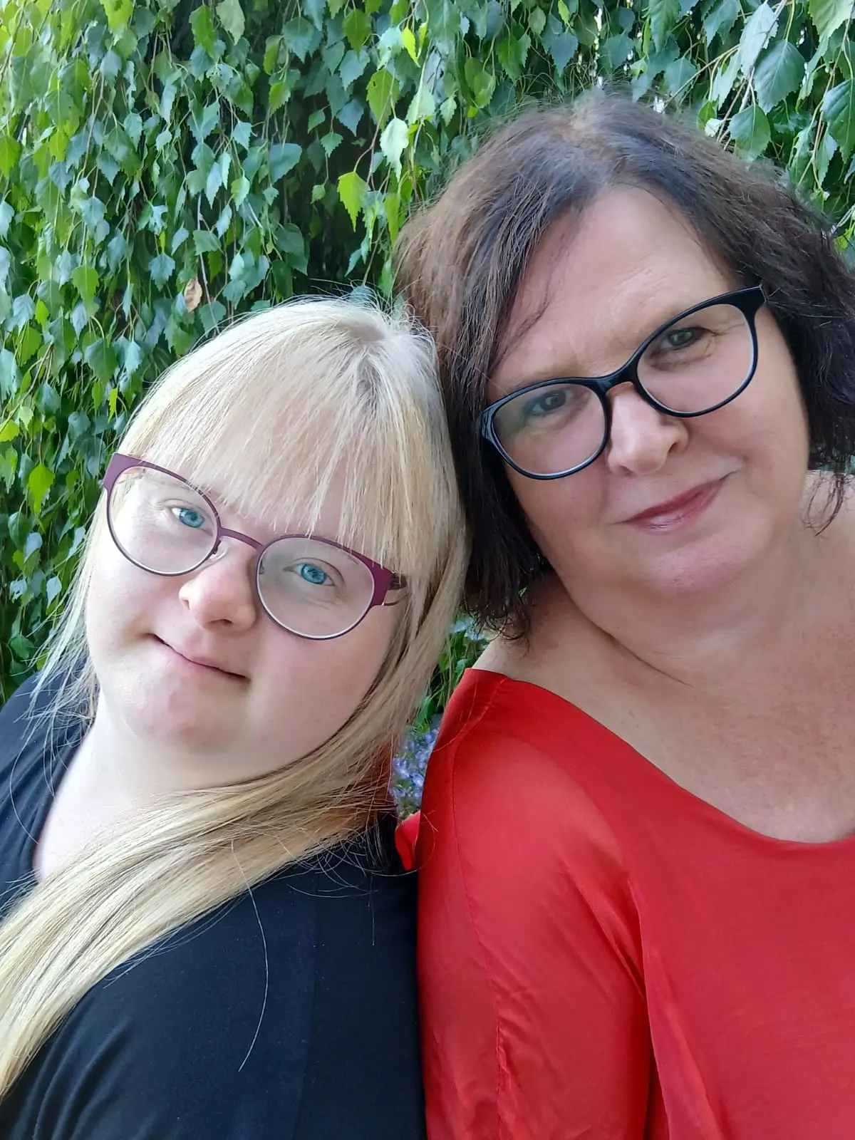 Zwei Frauen mit Brille lächeln vor grüner Pflanzenwand im Freien