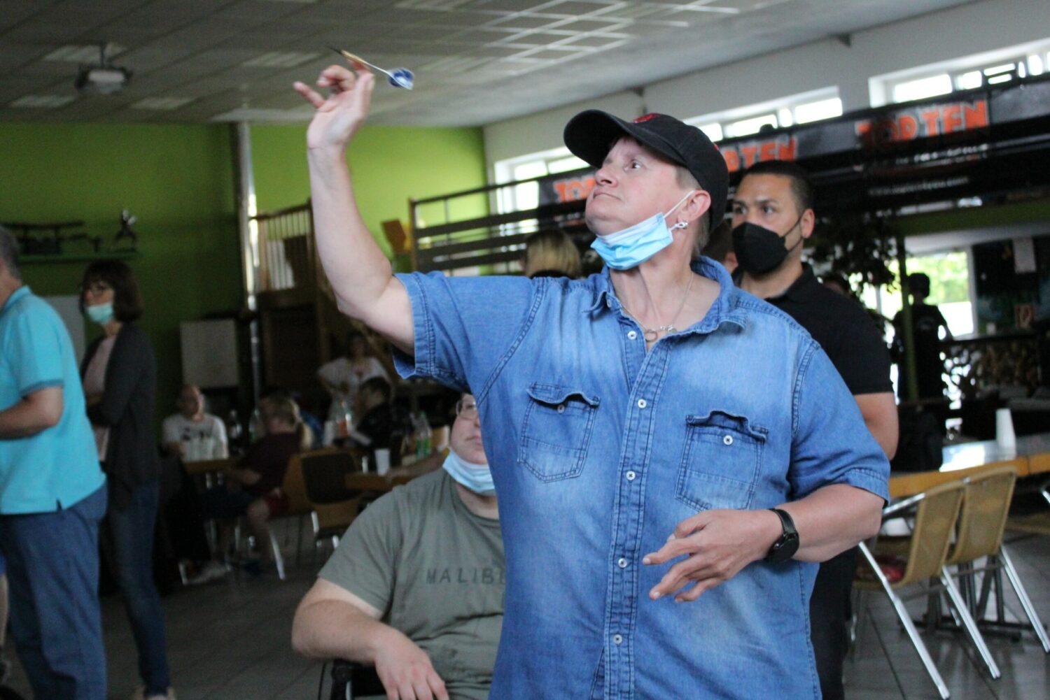 Mann wirft Dartpfeil im Clubraum, Zuschauer beim Darts-Turnier im Hintergrund.