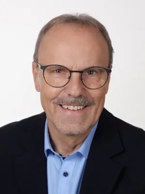 Porträt von Dirk Botzon mit Brille, Schnurrbart, Anzugjacke und blauem Hemd vor weißem Hintergrund.