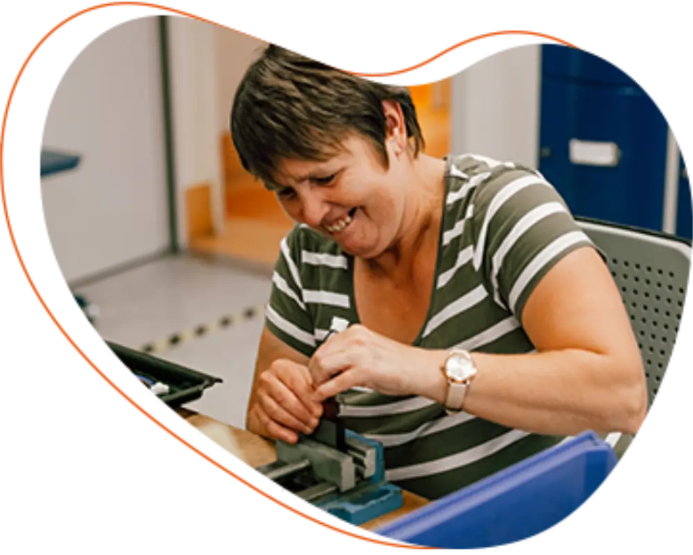 Frau in Werkstatt arbeitet am Schraubstock, montiert Bauteil mit Werkzeug, lächelt konzentriert.