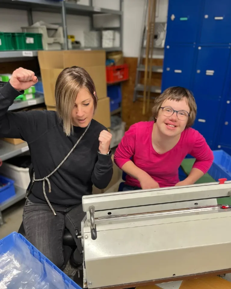 Zwei Frauen freuen sich bei Teamarbeit in der Werkstatt neben einer Verpackungsmaschine.