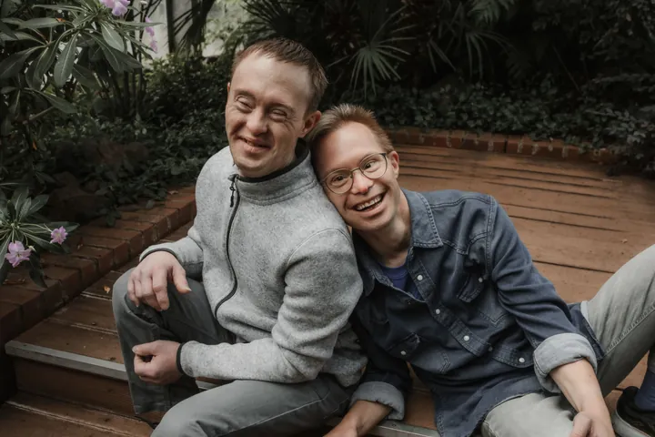 Zwei Freunde sitzen lächelnd auf Holzstufen im grünen Garten, Kopf an Schulter.