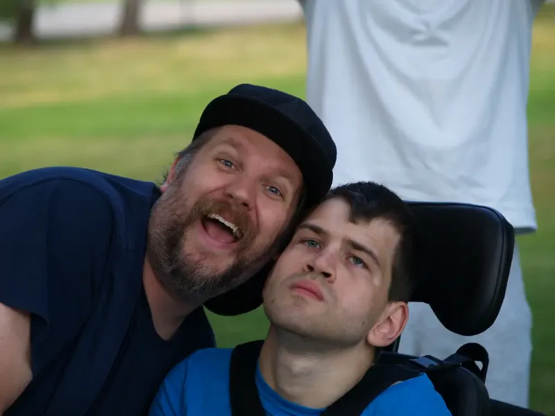 Zwei Freunde im Park, Mann im Rollstuhl, lächelnder Begleiter, Porträt im Grünen.