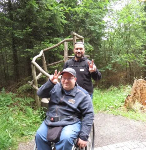 Zwei Freunde zeigen Peace-Zeichen im Wald, einer im Rollstuhl, auf Naturweg.
