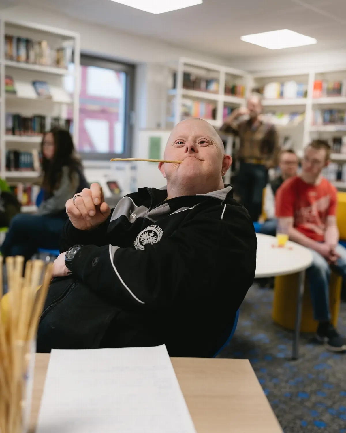 Mann mit Down-Syndrom lacht mit Snack am Tisch in der Bibliothek, Gruppenrunde im Hintergrund.