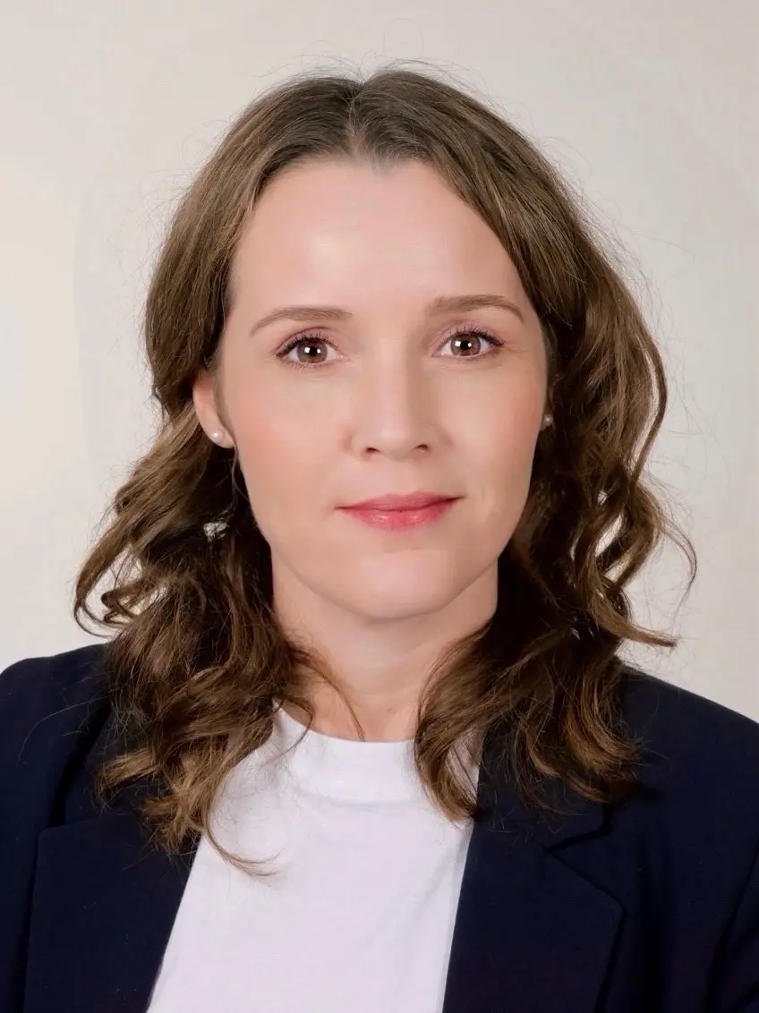 Professionelles Business-Foto: Frau mit braunen Locken im dunklen Blazer vor hellem Hintergrund.