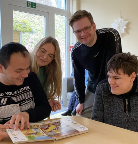 Vier junge Erwachsene lesen gemeinsam ein Buch am Tisch im Wohnzimmer.