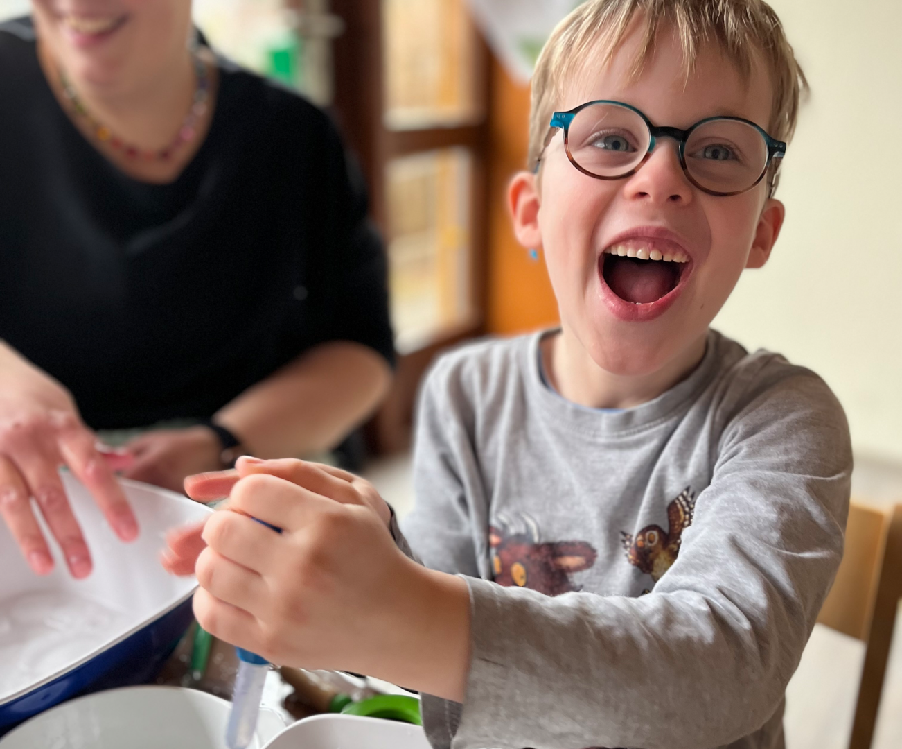 Lachender Junge mit Brille rührt Teig in Schüssel, Mutter hilft in der Küche.