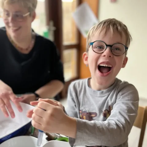 Lachender Junge mit blauer Brille rührt am Tisch, Oma lächelt im Hintergrund.