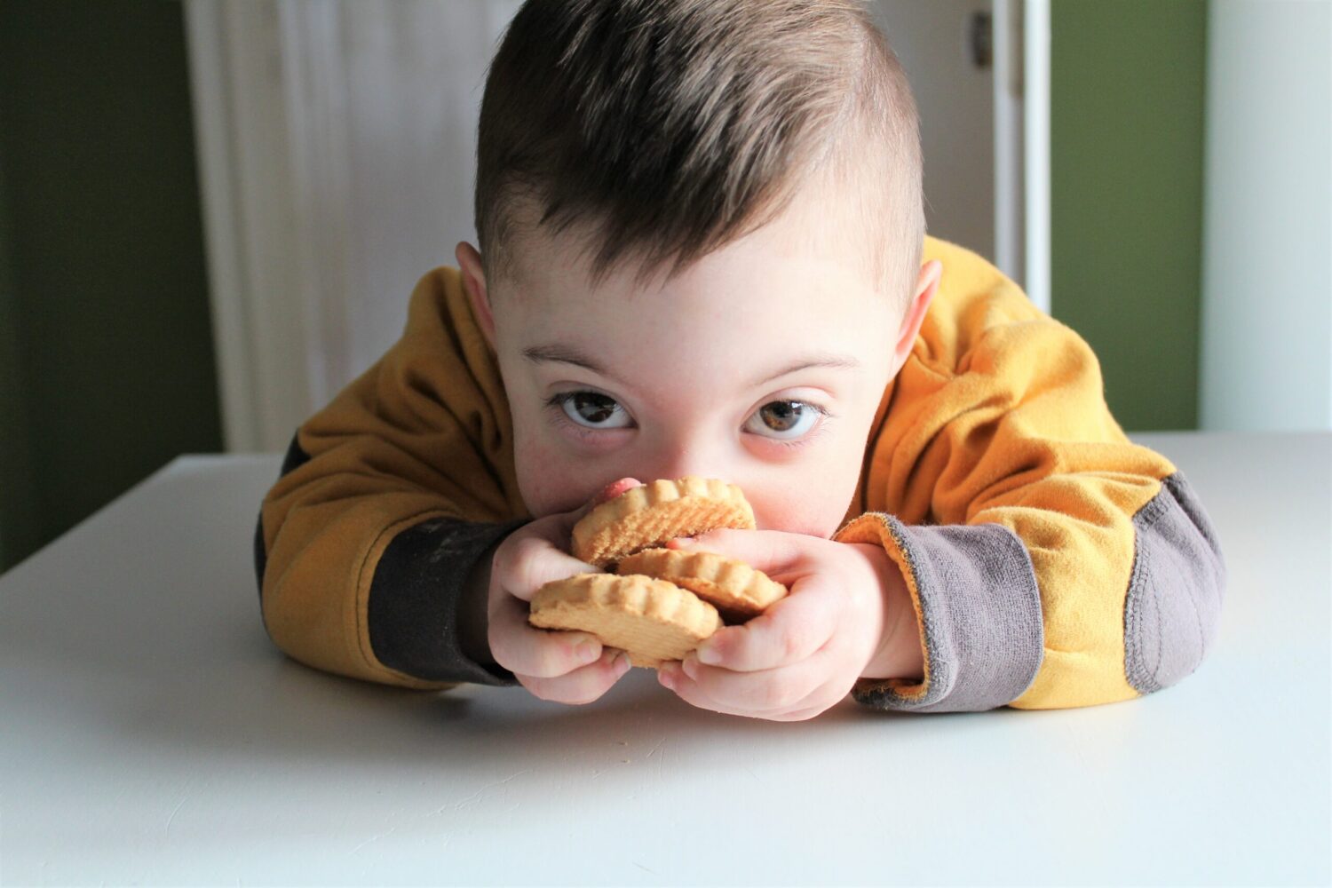 Junge am Tisch hält zwei Kekse und schaut neugierig in die Kamera.