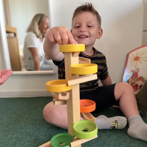 Junge spielt begeistert mit bunter Murmelbahn aus Holz, Mutter beobachtet im Hintergrund.