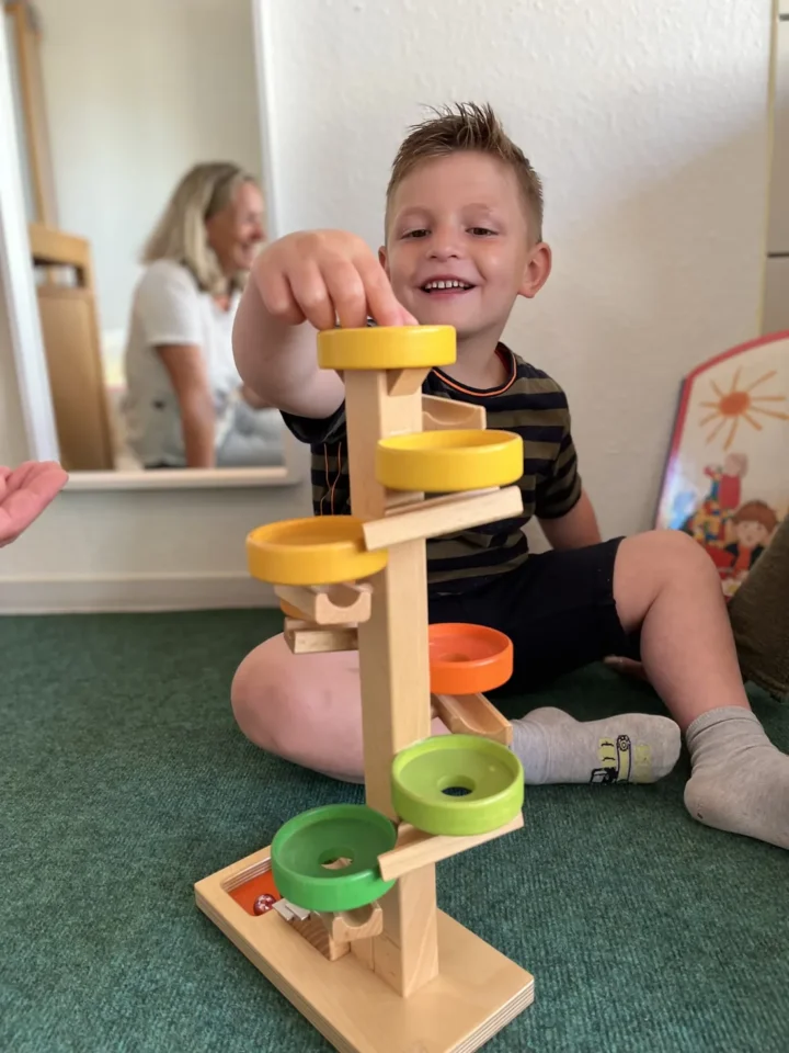 Junge spielt lächelnd mit Holz-Murmelbahn auf dem Wohnzimmerteppich, Mutter schaut zu.