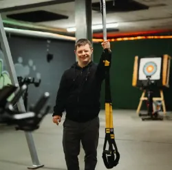 Junger Mann trainiert mit TRX Suspension Trainer im Fitnessstudio und lächelt.