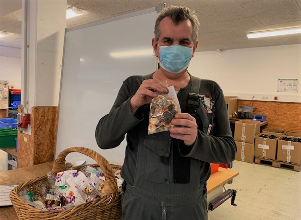 Mann mit Maske zeigt Plätzchentüte in Werkstatt Eibelshausen, Korb voller Plätzchentüten.