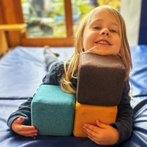 Fröhliches Kind auf blauer Spielmatte umarmt bunte Schaumstoffwürfel im Kinderzimmer.