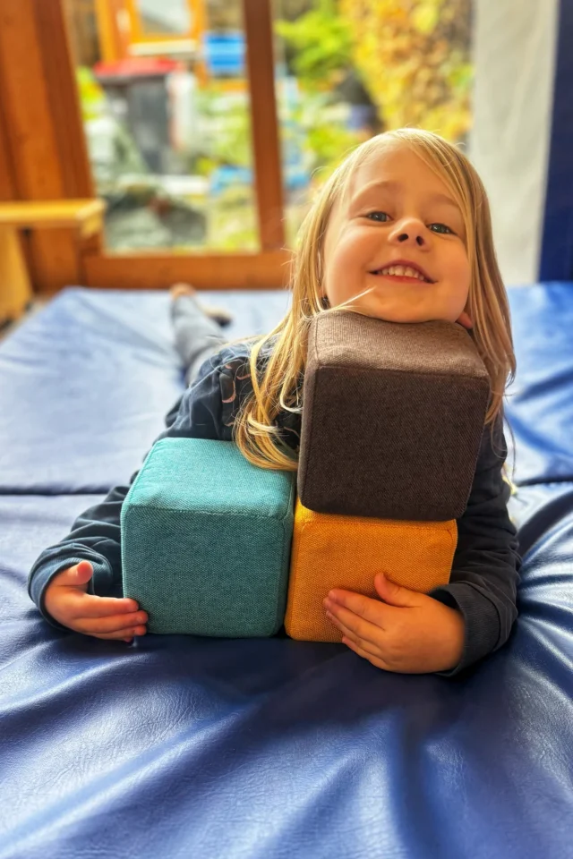 Lachendes Kind auf blauer Spielmatte mit gestapelten, bunten Softwürfeln im Kinderzimmer.