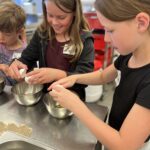 Drei Kinder im Kochkurs schlagen Eier in Schüsseln auf einem Edelstahltisch auf.