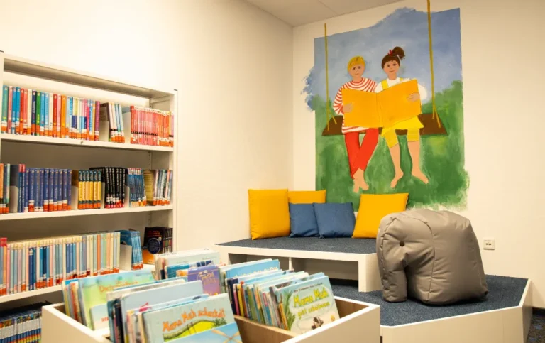 Heller Kinderlesebereich mit Bücherregalen, Sitzkissen und Wandbild in der Bibliothek.