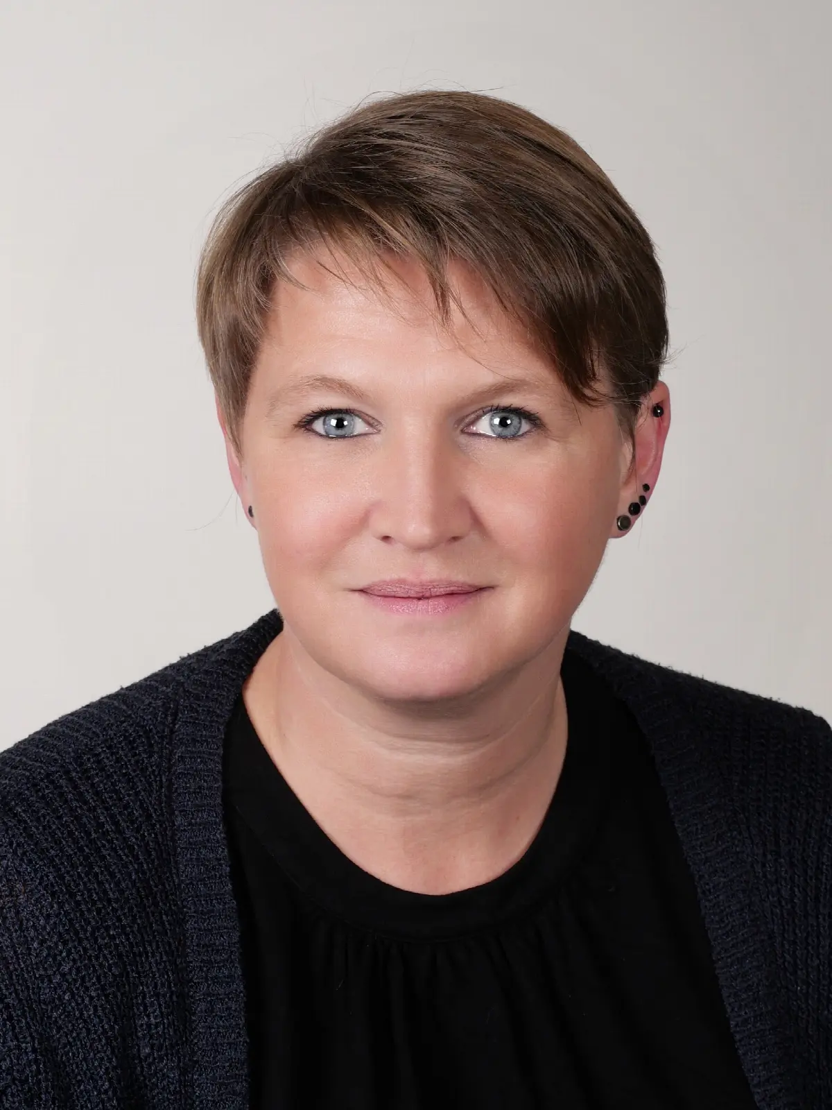 Professionelles Porträt einer Frau mit kurzen braunen Haaren und Ohrpiercings vor hellem Hintergrund.