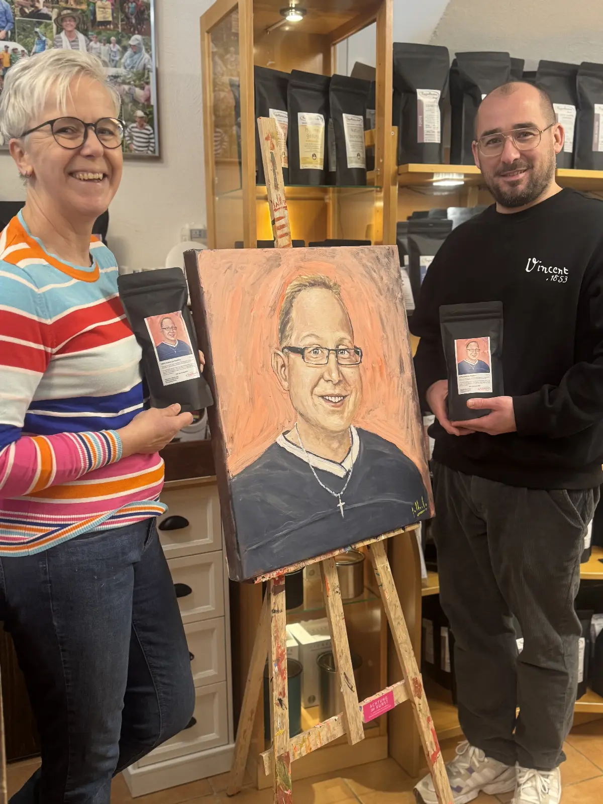 Zwei Personen präsentieren Porträt auf Staffelei im Kaffeeladen und halten Kaffeebohnen-Tüten.