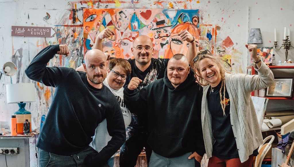 Fünf Künstler im Atelier posieren im Strongman-Stil vor bunt-abstraktem Gemälde.