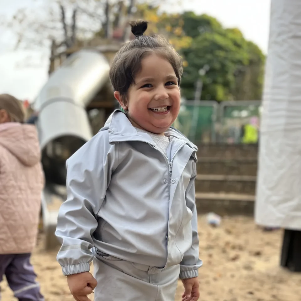 Lachendes Kind im grauen Regenanzug mit Gummistiefeln auf sandigem Spielplatz.