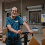 Lagerarbeiter mit Hubwagen lächelt im Versandlager vor Paletten und Kartons.