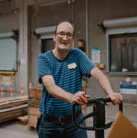 Lagerarbeiter mit Hubwagen lächelt im modernen Lagerhaus zwischen Paletten und Kartons.