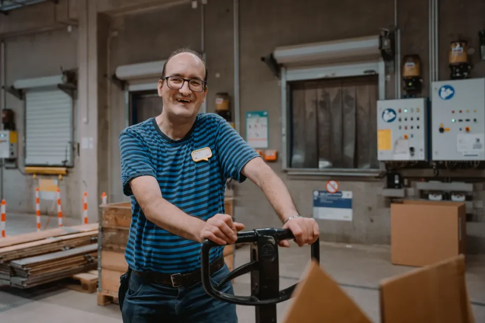 Freundlicher Lagerarbeiter mit Handhubwagen in sauberem Lager zwischen Paletten und Kartons.