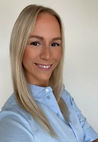 Professionelles Business-Porträt einer lächelnden blonden Frau im hellblauen Hemd vor neutralem Hintergrund.