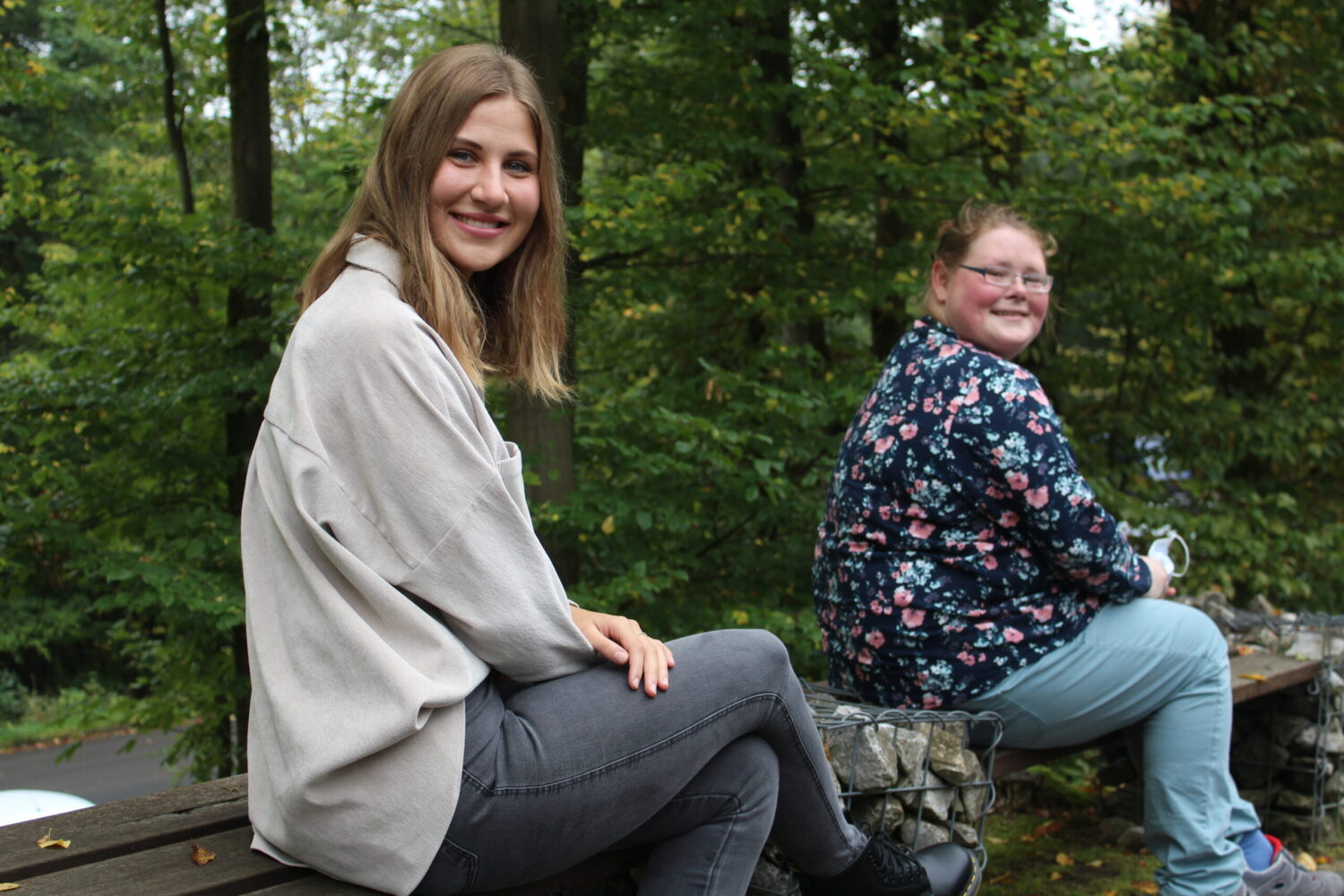 Lea Wölke und Lisa Benner lächeln im Wald auf Bank und Steinmauer.