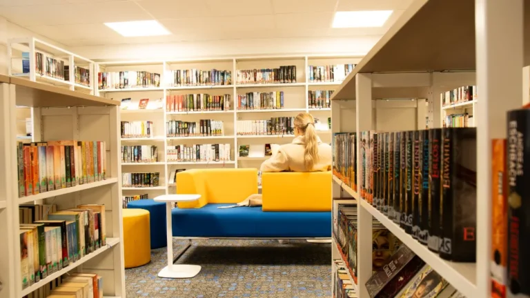 Helle Stadtbücherei: Frau liest auf blau-gelbem Sofa zwischen weißen Bücherregalen.