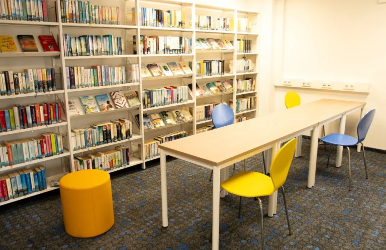 Moderner Bibliotheks-Lesebereich mit langem Tisch, bunten Stühlen und vollen Bücherregalen.