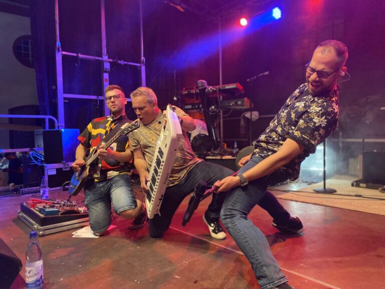 Drei Musiker bei explosivem Live-Auftritt mit Keytar, Gitarre und Mikrofon auf Bühne.