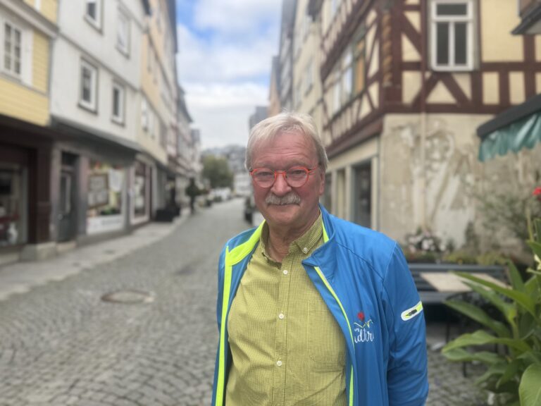 Porträt von Lothar Klein mit roter Brille in historischer Altstadt auf Kopfsteinpflaster.