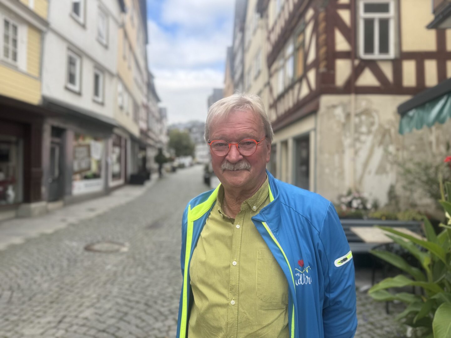 Porträt von Lothar Klein mit roter Brille in historischer Altstadt auf Kopfsteinpflaster.