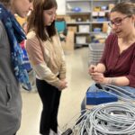 Mädchen lernt Kabelmontage in der Technikwerkstatt, Frau erklärt Steckverbinder am Arbeitstisch.