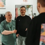 Drei Männer im Kreativstudio vor Spiegel, Modeatelier mit Kleidung und Pflanzen im Hintergrund.