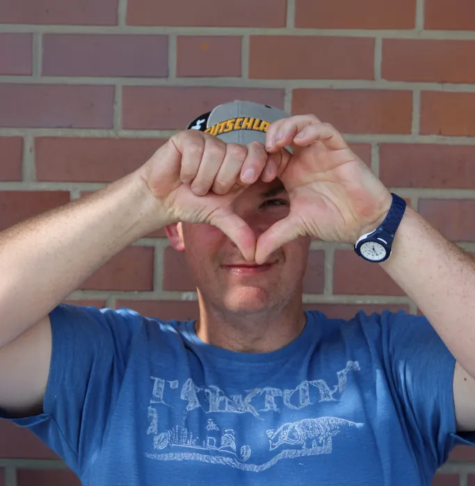 Mann mit Cap formt Herzzeichen mit Händen vor Ziegelwand, Outdoor Porträt.