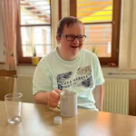 Fröhlicher Mann mit Brille trinkt Kaffee am Holztisch in heller Cafeteria.