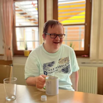 Fröhlicher Mann mit Brille am Holztisch hält Kaffeetasse im hellen Wohnzimmer.