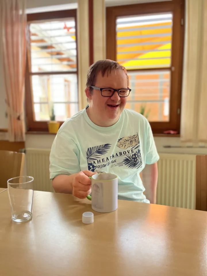 Fröhlicher Mann mit Brille trinkt Kaffee am Holztisch in heller Cafeteria.